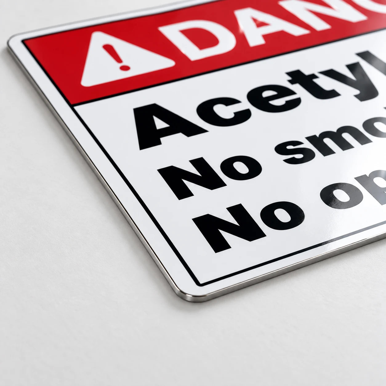 A close-up of a rectangular warning sign with a red header, white background, black text reading 'DANGER Acetylno smok No op', and rounded metal corners on a light gray surface.