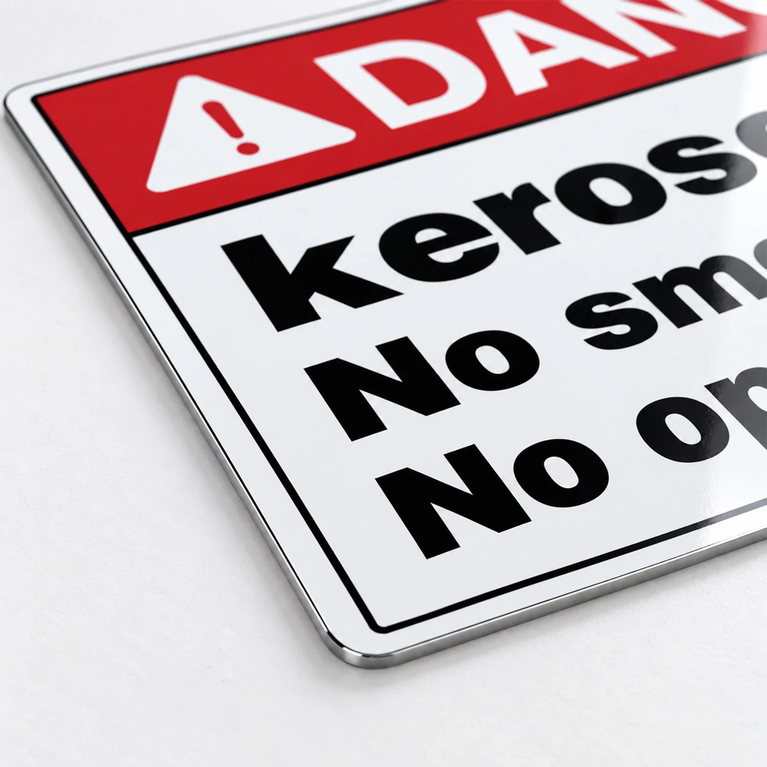 A close-up of a square warning sign with a red header and white background, reading 'DANGER keros No sm No op' in black text, photographed on a light neutral surface.