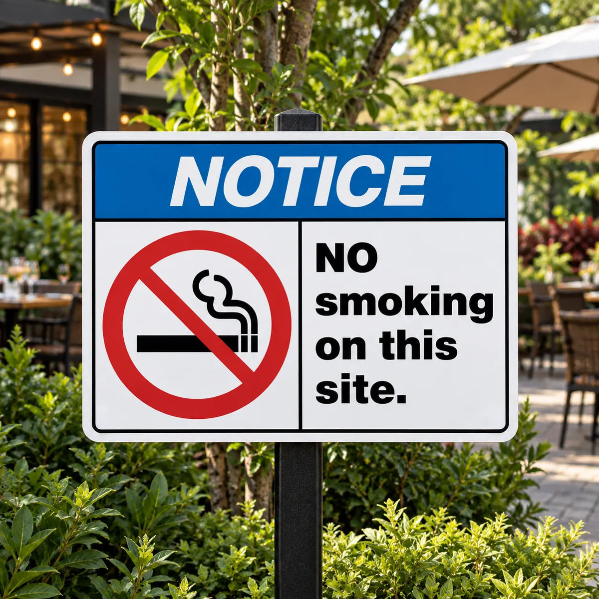 A rectangular outdoor smoking prohibition sign on a black post, with a blue header reading 'NOTICE' and a red no-smoking symbol alongside the text 'NO smoking on this site.' The sign is photographed in a real patio/garden environment with plants and seating in the background.