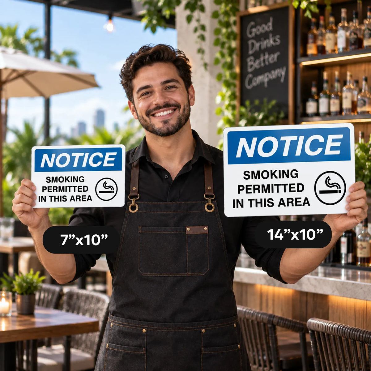 A smiling man in a black apron stands in a restaurant holding two smoking notice signs, one 7"x10" and one 14"x10", against a blurred bar and patio background.