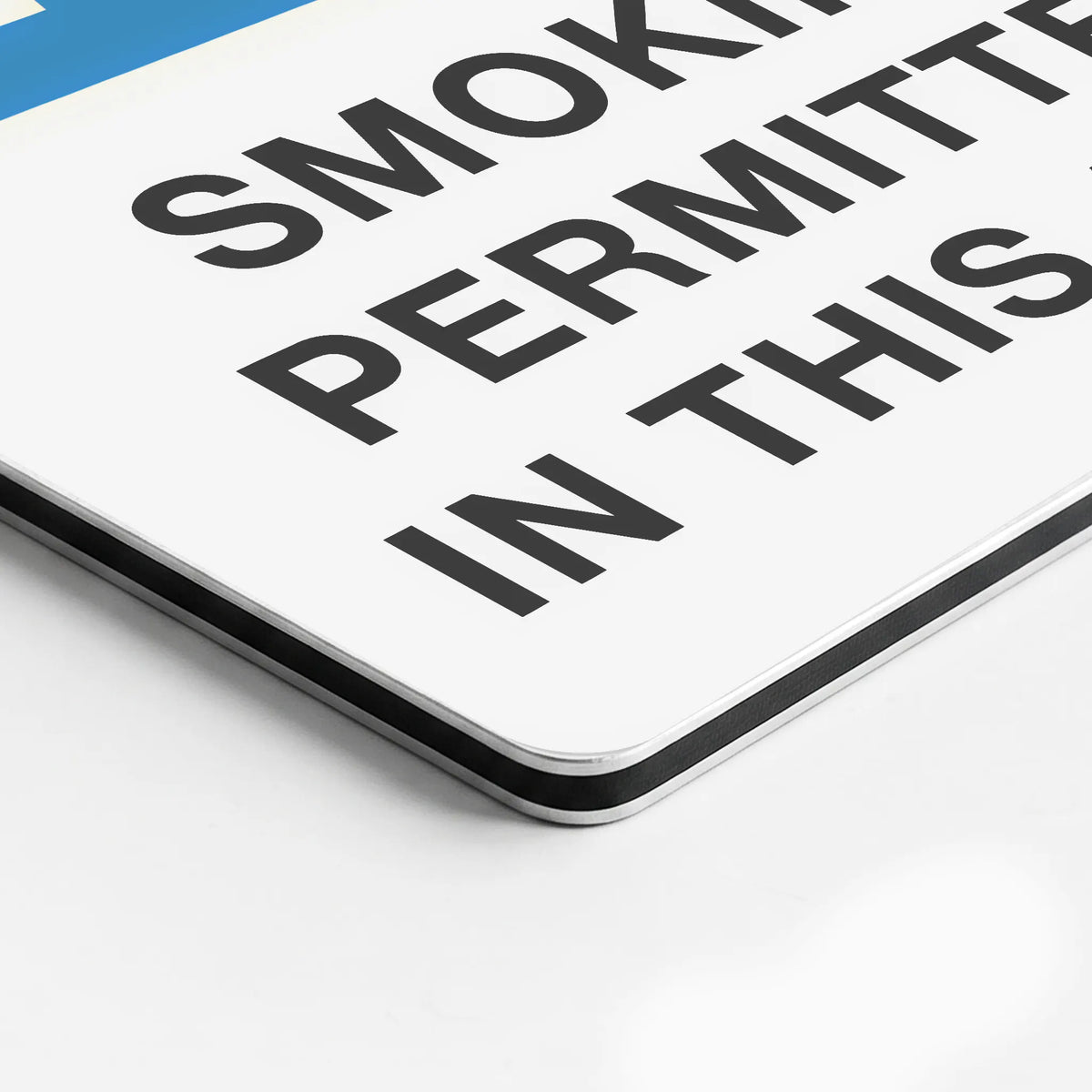 A close-up of a white rectangular sign with bold black text reading 'SMOKING PERMITTED IN THIS AREA' and a small blue section in the top-left corner, photographed on a white surface.