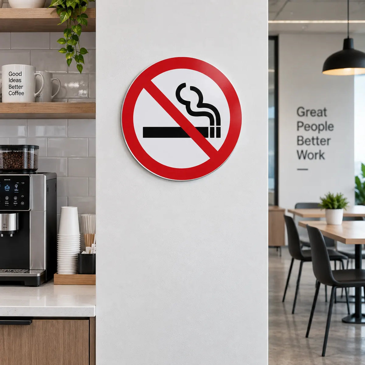 A circular 'No Smoking' sign with a red border and slash over a black cigarette icon, mounted on a white wall in a modern office and kitchen environment.