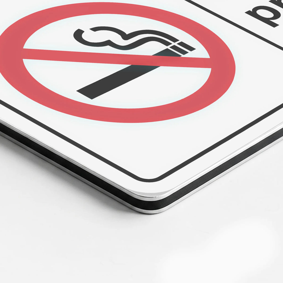 Close-up of a square white sign with a red no-smoking symbol and black border, photographed on a light gray background.