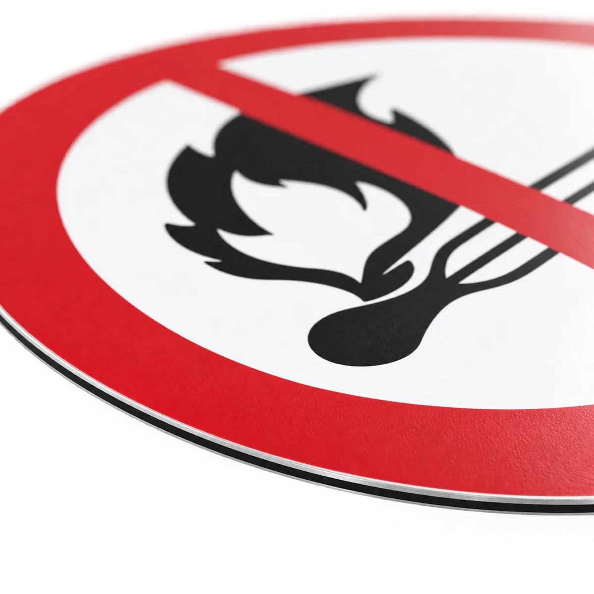 A close-up of a circular fire prohibition sign with a red border, white center, black flame-and-match icon, and a red diagonal slash, photographed on a white background.