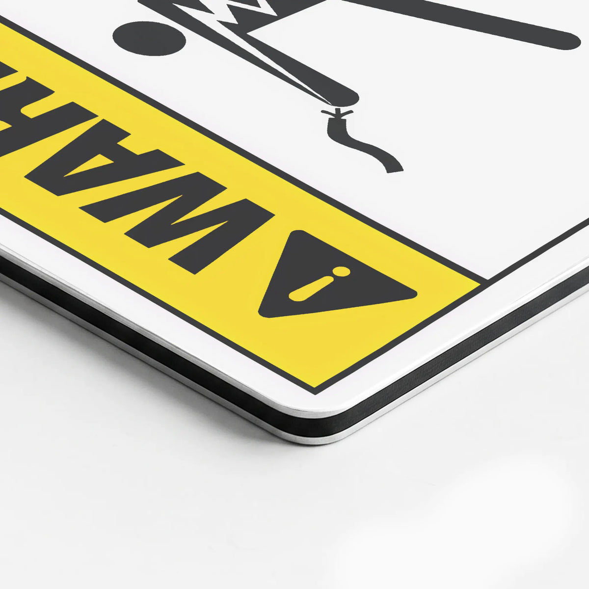 A close-up of a warning sign with a yellow background, black border, black warning symbol, and partially visible bold text on a white surface.