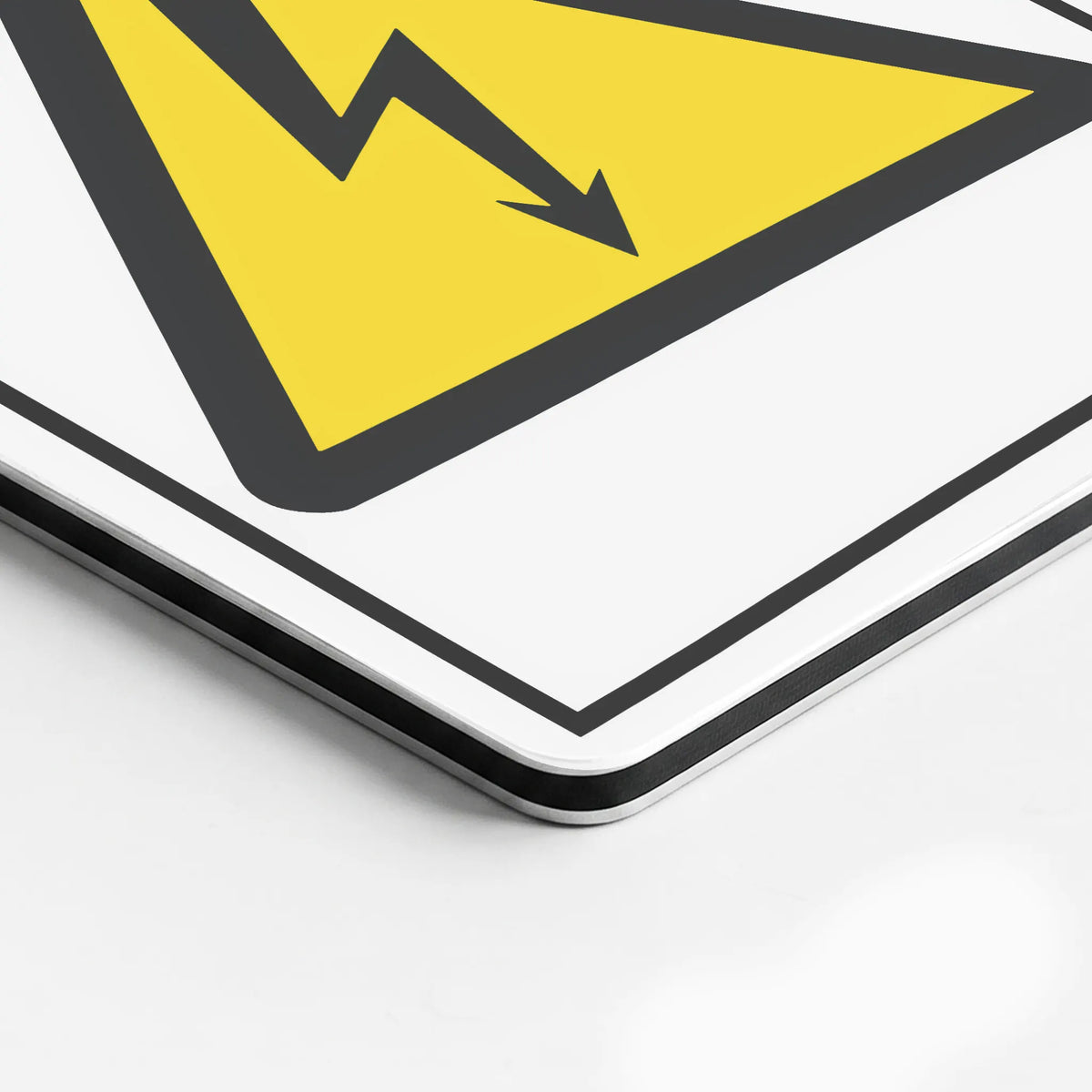 A close-up of a square warning sign with a yellow triangle, black lightning bolt, and black border on a white background, shown at an angle on a light surface.